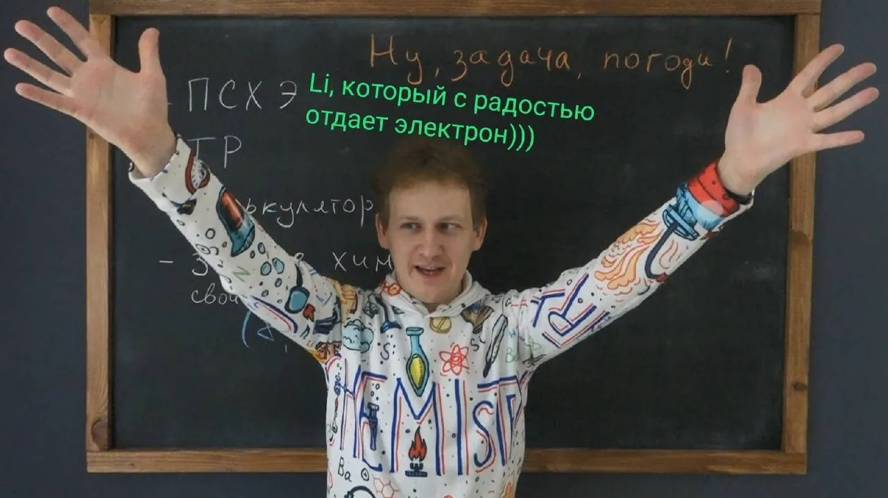 Александр Евсюков в толстовке с химической росписью развёл руки у школьной доски, на ней мелом написано «Ну, задача, погоди!»
